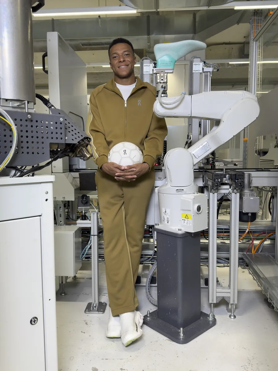 Kylian Mbappé debout en survêtement kaki, un ballon blanc dans les mains, à côté d’un bras robotisé portant une forme de chaussure turquoise