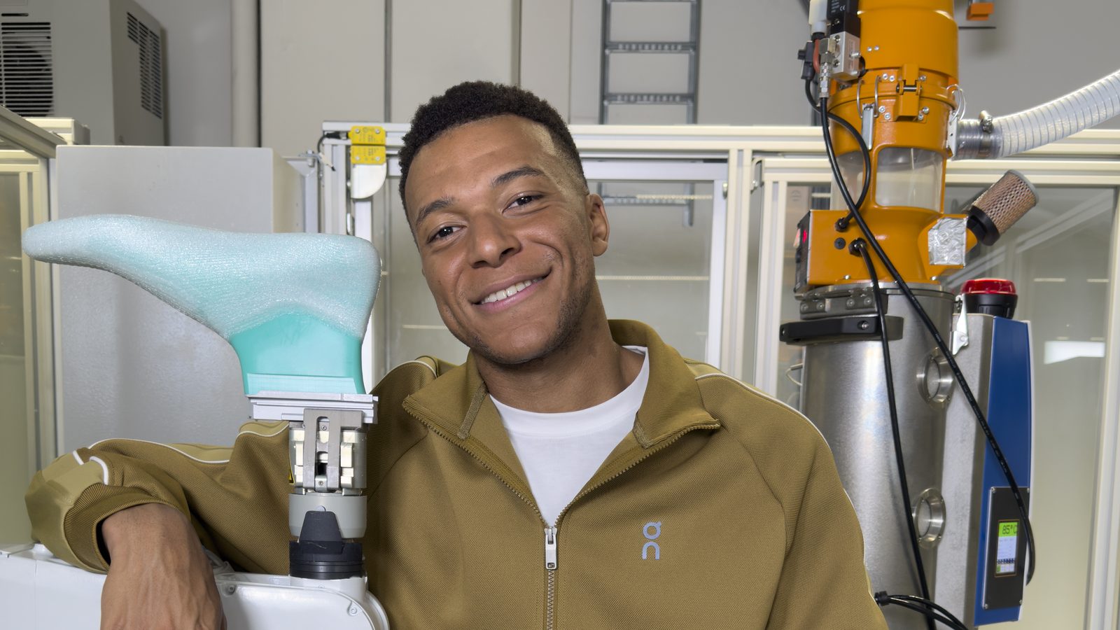 Kylian Mbappé souriant, en survêtement On kaki, accoudé à un bras robotisé qui tient une forme de chaussure, dans un atelier des On Labs à Zurich