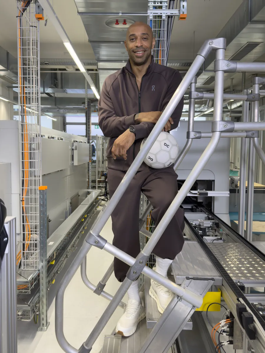 Thierry Henry assis sur un escalier métallique dans un atelier, en survêtement marron, un ballon blanc sous le bras